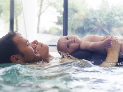 Moment de complicité en famille dans une piscine ou un bain thermal Valdys. Un père et une mère sont allongés dans l'eau claire avec leur bébé qui regarde curieusement vers l'objectif, dans une ambiance lumineuse et relaxante